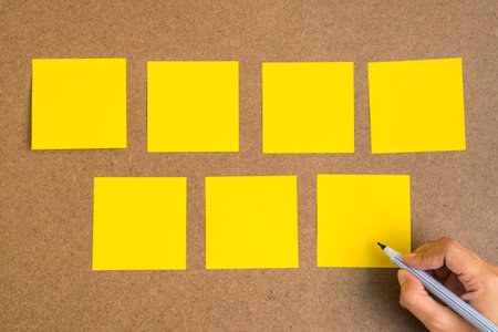 Seven blank yellow sticky notes on brown wood board with human hand hold a pen