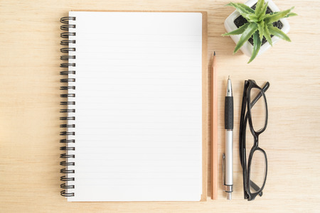Desk above, Top view of open spiral notebook with brown pencil, pen, black eye glasses and small plant pot on wooden backgroundの写真素材