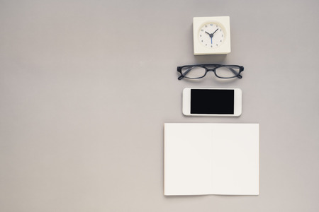 Desk above, table top view of office desk with notebook, mobile phone, clock, eyeglasses on grey backgroundの写真素材
