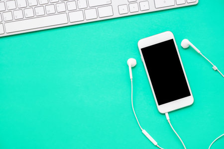 Top view of mobile phone with headphone and white keyboard on fresh green background