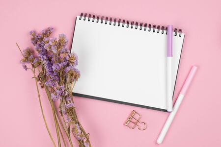 Top view of blank empty notebook with pens and paper clip and violet statice flower bouquet on pastel pink backgroundの写真素材