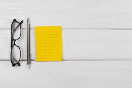 Top view of a yellow blank sticky note with pencil and eyeglasses on white wooden backgroundの写真素材