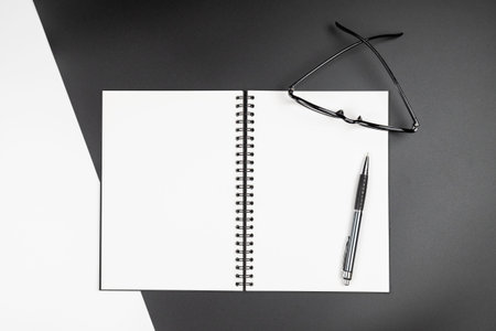 Top view of an empty open notebook with black pencil and eyeglasses on black and white background