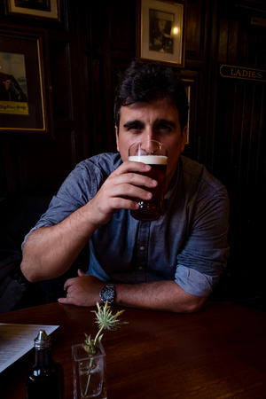 Brazilian man holding pint of dark beer and taking a sip in an traditional English pubの写真素材