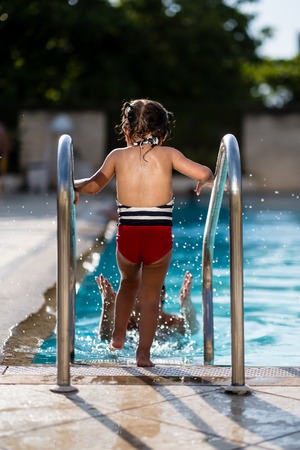 Little girl entering swimming pool via metalic stairs during a sunny day in summer, while mother splashes water on herの写真素材