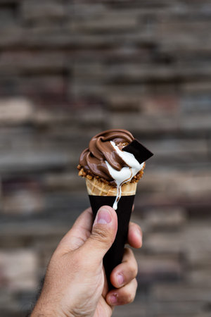 Man holding melting vanilla and chocolate ice cream cone in hand, peanuts around cone, very blurred background, small depth of field. Dark background.の写真素材