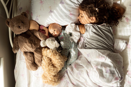small toddler girl sleeping with teddy bears and dolls on small bed, beautiful light shining through...
