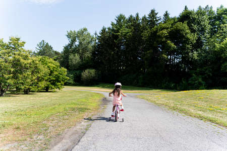 Young girl in pink dress and helmet riding pink bike on a park sidewalk, seen from behindの写真素材