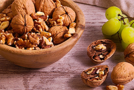 45 degrees photo of group of walnuts into wood bowl and some walnuts and a group of green grapes beside, all over wood board and brown path at the backの写真素材