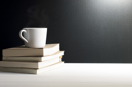 Still life - A cup of hot coffee put on old books on white wooden table in font of grunge black background.の写真素材