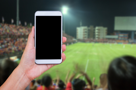 Hand holding smartphone with soccer screen, blurred football stadium background.の写真素材