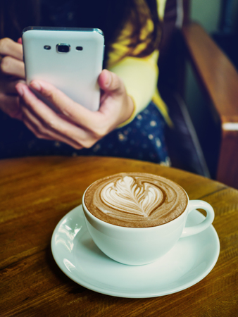 Latte coffee cup on wood table with woman holding smartphone in cafe. Vintage color tone styleの写真素材