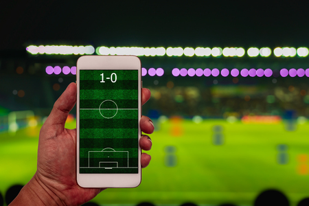 Hand holding smartphone with football field on blur soccer stadium background.の写真素材