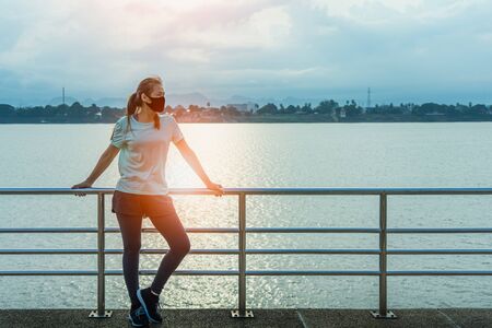 Young fitness Asian woman standing resting tired with wearing a protective mask after running and jogging an outdoor workout on the riverwalk in the morning for lifestyle health.の写真素材