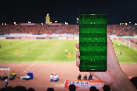 Hand holding smartphone with blur soccer field stadium background.の写真素材