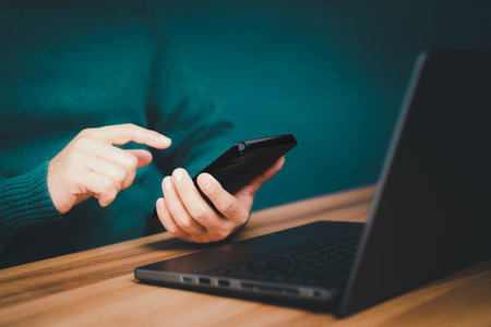 Male hand using a smartphone while working on computer laptop at office for investment analyzing company financial report balance with mobile phone. Concept of people are working with mobile devices.の写真素材