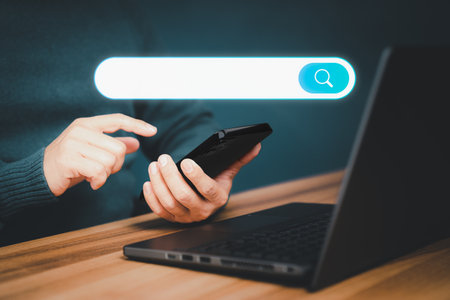 Adult man hand working with a mobile phone and computer laptop on the desk. Searching browsing internet data information with blank search bar. Search Engine Optimization SEO networking concept.の写真素材