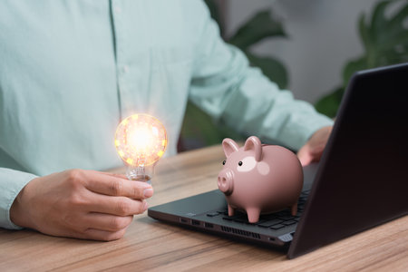Man holding a light bulb while working on a computer to think and generate new ideas for business profitability with saving money, sales and purchases in online stores internet shopping.の写真素材