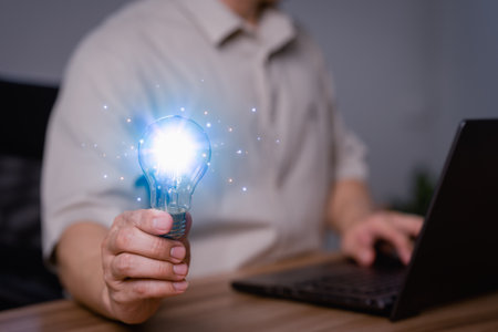 Adult man holding a light bulb while working on a computer laptop to think and generate new ideas through technological innovation and creativity. Idea inspired by online technology and innovation.の写真素材