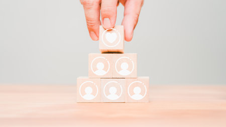 Hand put a wooden cube block with a heart icon on the top of stacking wooden cubes standing with employee or team icon. Business concept of teamwork, goal achievement, financial growth, target.の写真素材