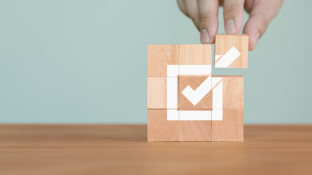 A hand places a wooden block to complete a large check mark made from smaller blocks. The image...