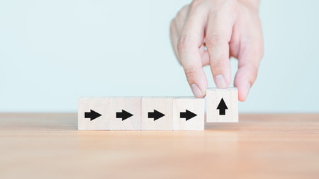 Hand arranging wooden cube blocks with arrows and one upward arrow, symbolizing change, growth, and innovation, placed on a wooden table against a light background.の写真素材