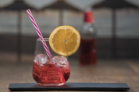 Photo detail of a tasty red summer cocktail served with ice cubes, a straw and a slice of orange on a black stone table, tequila bottles in the backgroundの写真素材