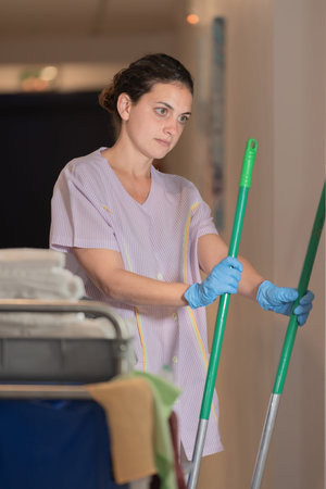 Thoughtful woman dressed in a white uniform, staring at a pair of brooms in the hallway of a clinic....