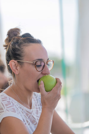 Face of woman eating juicy apple while looking down wearing glasses and white shirt with unfocused background. Vertical image.の写真素材