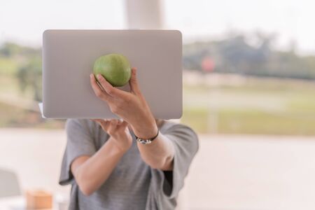 Girl whose face is not seen wearing a gray shirt holding a laptop and placing an apple in front of the laptop with the background out of focus. Female beauty concept.の写真素材