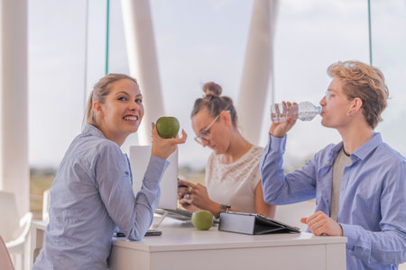 Attractive girl holding a green apple in one of her hands while sharing a table with two more people who use a laptop. Technology conceptの写真素材