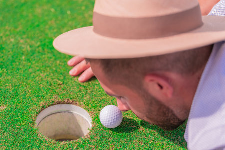 A young man with a beard and a hat, closely observing a golf ball on a green field. Concept of a day of relaxation and leisure. Top viewの写真素材