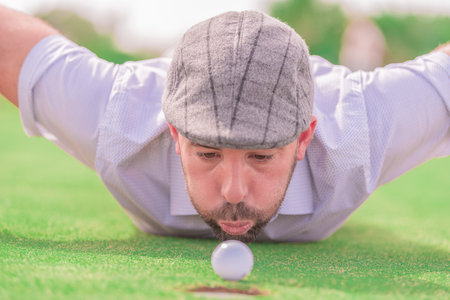 Close-up of the face of a man with a grey beard and cap, trying to move a white golf ball blowing, with the background out of focus.の写真素材