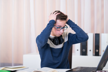 Overwhelmed teenage boy puts his hands to his head while watching a laptop screen on an out of focus...