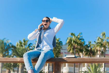 Confident looking woman raises her sunglasses and looks up while talking on her phone in a blue sky...