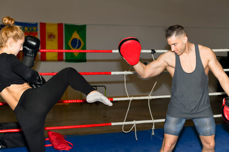 Confident looking woman in boxing gloves trains kicking against a focus pad being held by a male instructor in a boxing ring. Fitness concept.の写真素材