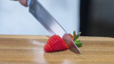 Close-up of a delicious looking fresh strawberry being but into slices with a sharp metal knife on a wooden board on an out of focus background. Healthy and vegan food concept.の写真素材