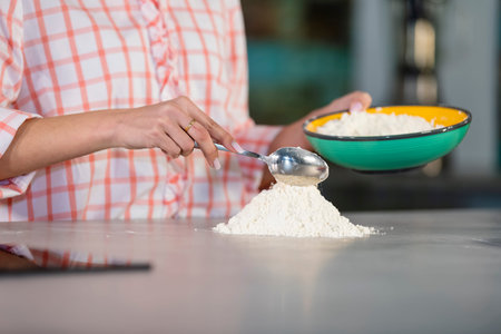 Close-up of a woman holding a bowl adding flour to a mound with a spoon on the kitchen counter on an out of focus background. Cooking concept.の写真素材