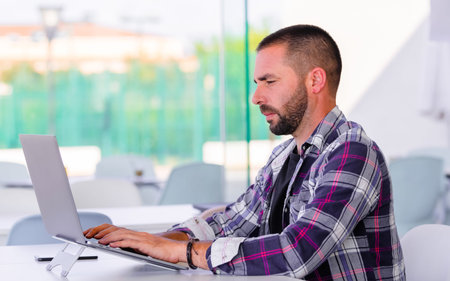 Close-up of a handsome young man sitting and typing on his laptop on a table on an out of focus...