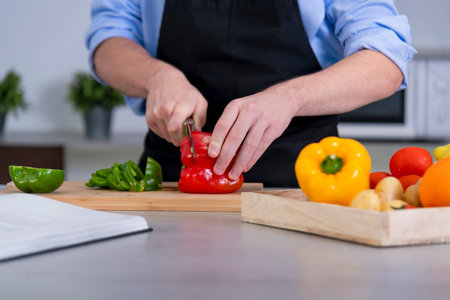 Close up of a young male cutting a red bell pepper on a cutting board surrounded by assorted vegetables on an out of focus background. Safety and cooking at home concept.の写真素材
