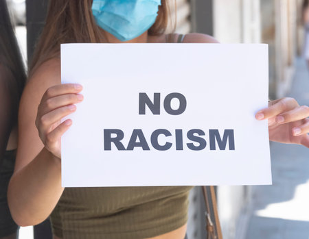 Close up of a young woman wearing a surgical mask holding a sign with a message on it on an out of...