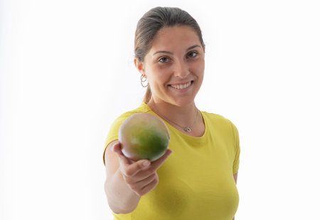 Close up of a smiling young beautiful woman offering an out of focus mango at the camera on a light background. Healthy food and lifestyle concept.の写真素材