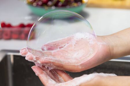 Close up of female hands holding a big soap bubble over an out of focus kitchen sink. Hygiene and fun concept.の写真素材