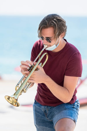 Handsome young man wearing glasses with his surgical mask pulled down playing a trumpet outdoors on...