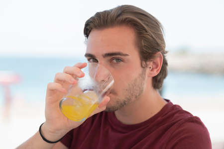 Close up of a handsome young man drinking orange juice while looking at the camera on an out of focus background. Leisure and healthy food concept.の写真素材