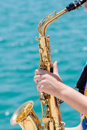 Close up of a shiny golden saxophone being held and played by a woman on an out of focus sea background. Street musician and lifestyle concept.の写真素材