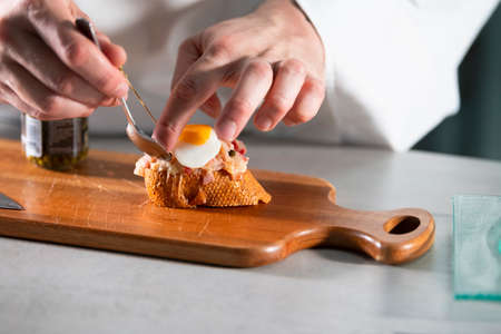 male hands using a spoon to place an egg on an open sandwich with assorted meat and cheeseの写真素材