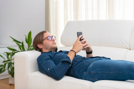 Close up of a handsome young man lying on a sofa and focused on his phone on an out of focus...