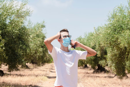 Young man wearing a surgical mask holding his head while talking on his phone on an out of focus...