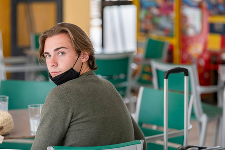 Young man wearing a face mask and a protective suit waiting at a train station while looking at the...